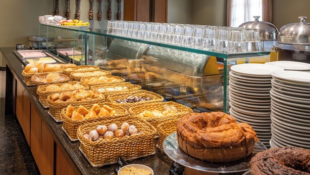 A buffet with various baked goods, including bread, pastries, and cakes, displayed on a counter with a glass case separating the food from the guests. - DELPLAZA Marabá São Paulo (São Paulo)