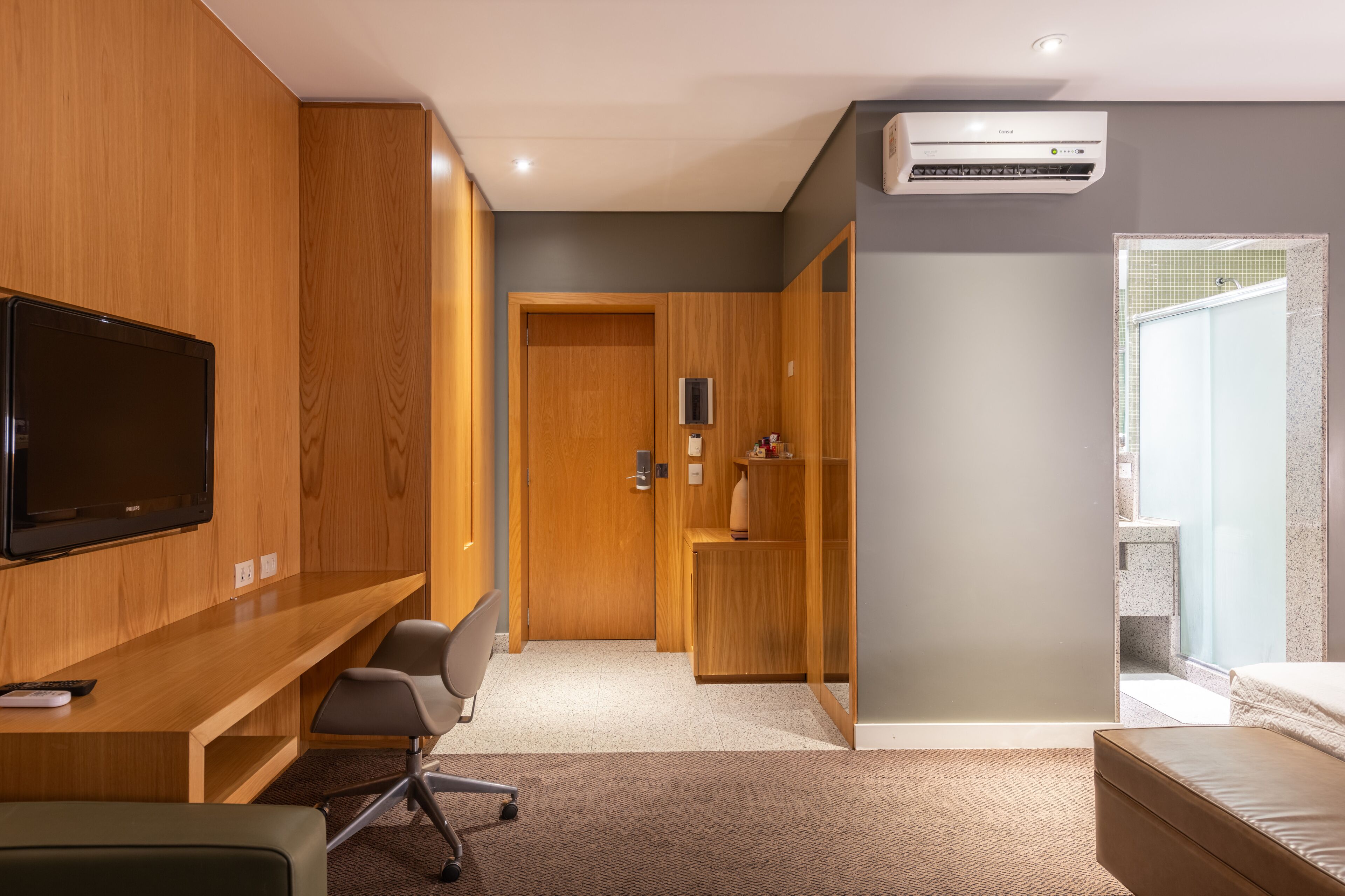A modern hotel room with a wooden desk, a television, a seating area, a wardrobe, and a bathroom with a shower and a mirror.