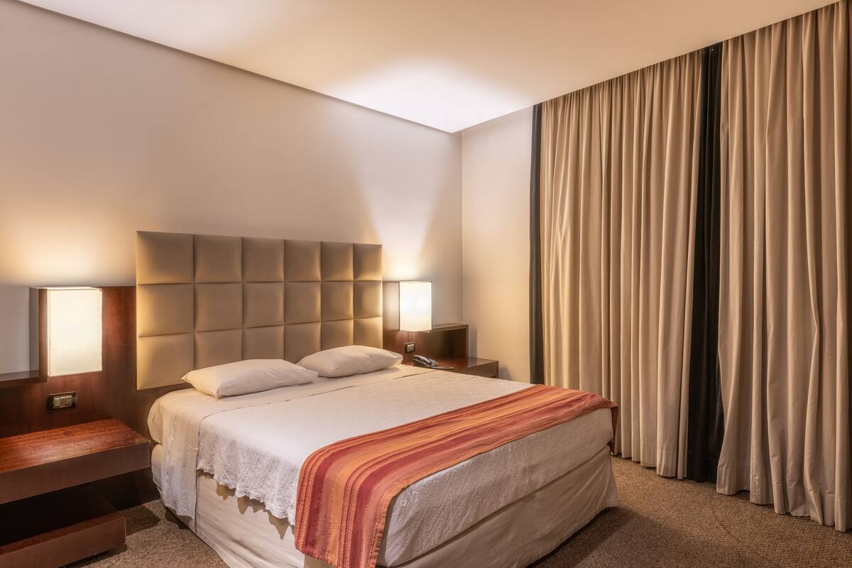 A hotel room with a large bed, a headboard with integrated lighting, bedside tables, and a view of a curtain.