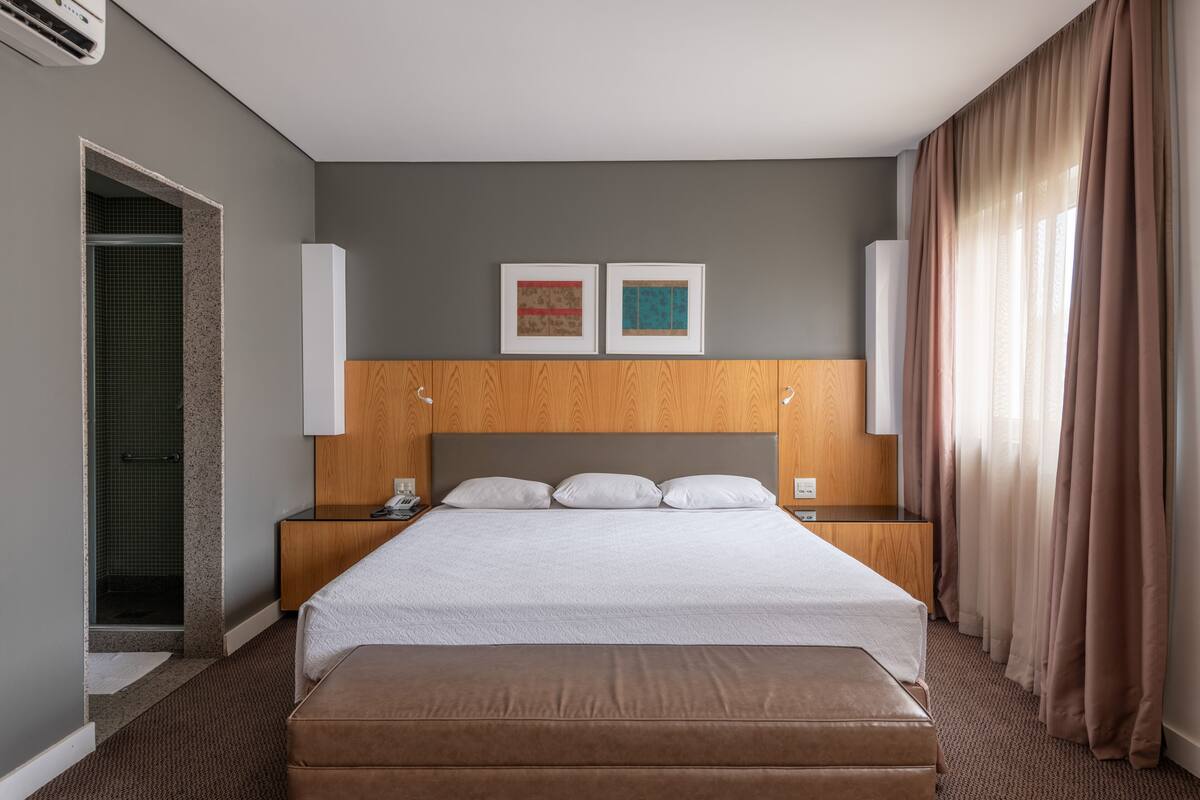 A hotel room with a large bed, wooden headboard, two bedside tables, a nightstand, a wooden bench, a shower area, and a window with curtains.