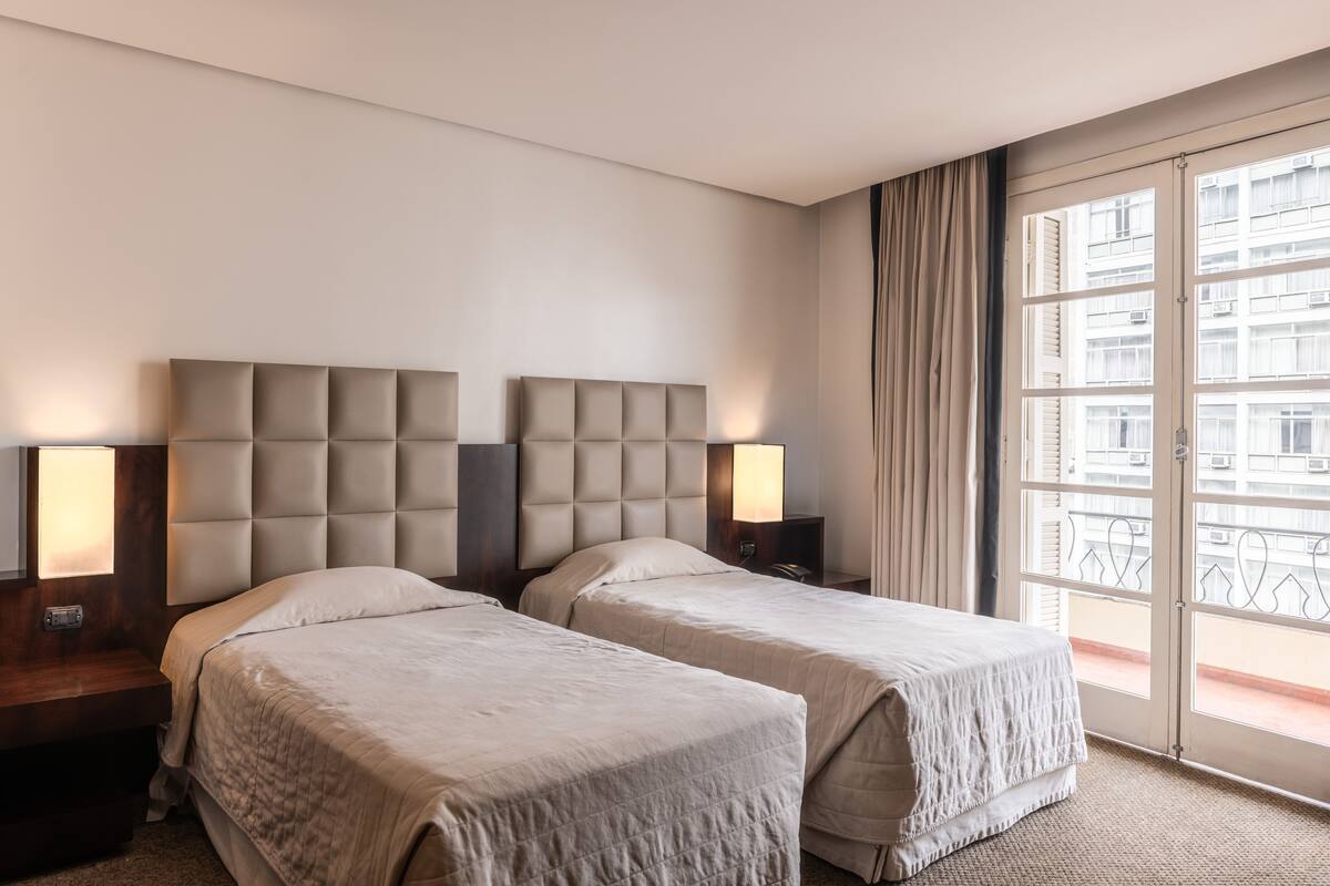 A hotel room with two beds, a large window with curtains, and a view of buildings outside.