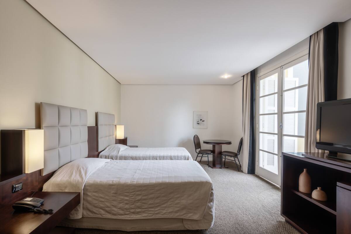 A hotel room with two beds, a desk with a telephone, a chair, a small table, and a television.