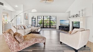 Ein geräumiges Wohnzimmer mit einer großen Couch, einem Relaxsessel und einem Fernseher. Es gibt einen Deckenventilator, einen Kamin und ein großes Fenster mit Blick auf Grünflächen.
