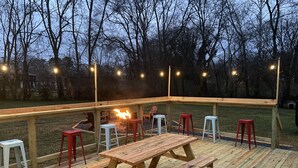 Una terraza de madera con una mesa de picnic y taburetes rojos y blancos, rodeada por una barandilla de madera y luces colgantes.