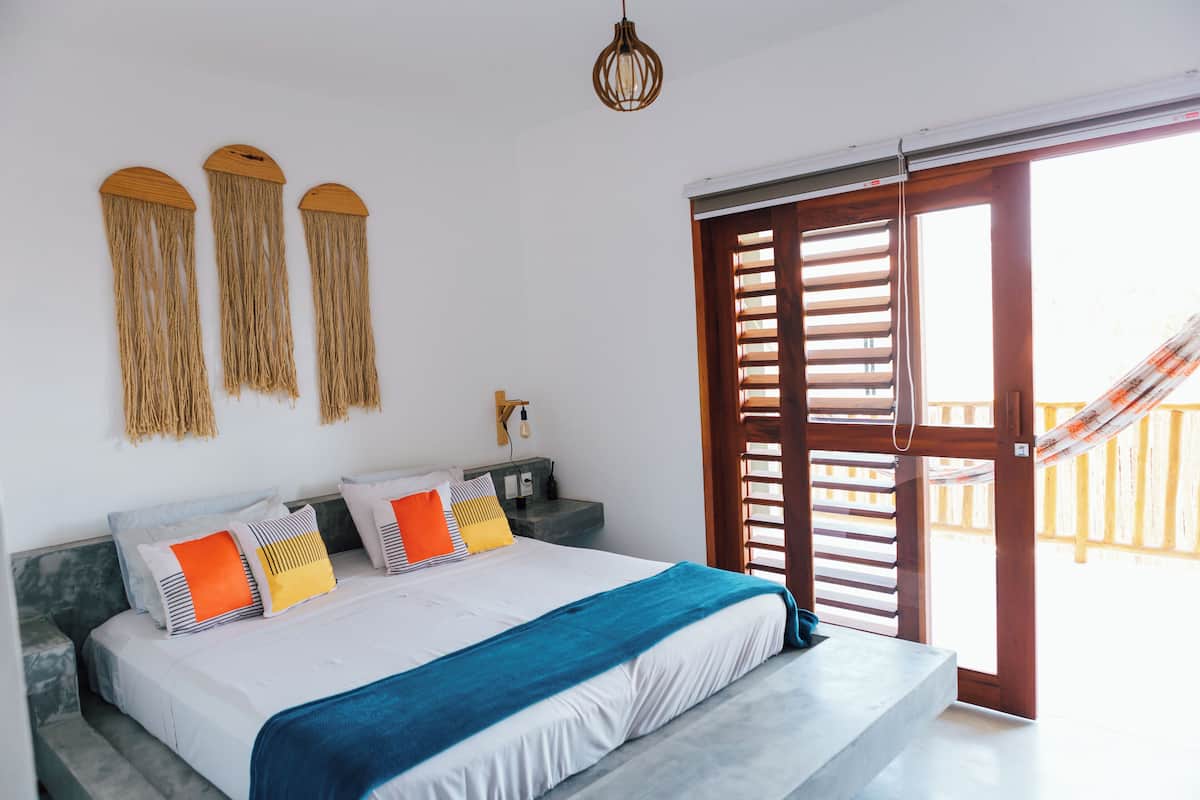 A bedroom with a bed, nightstands, a hanging light fixture, and a view of the outside through a wooden door.