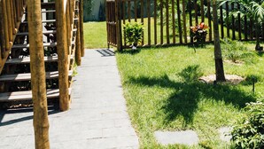 A wooden deck with a thatched roof, a stone pathway, and a fenced garden area with palm trees and shrubs. - Vila Tuga (Cruz)