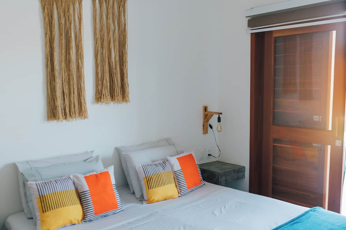 A bedroom with a bed, pillows, a bedside table, a wall-mounted light fixture, and a door.