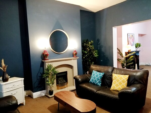 A living room with a fireplace, a leather sofa with patterned cushions, a wooden coffee table, and a white side table with decorative items. - Grand Palace in Gateshead (Gateshead)