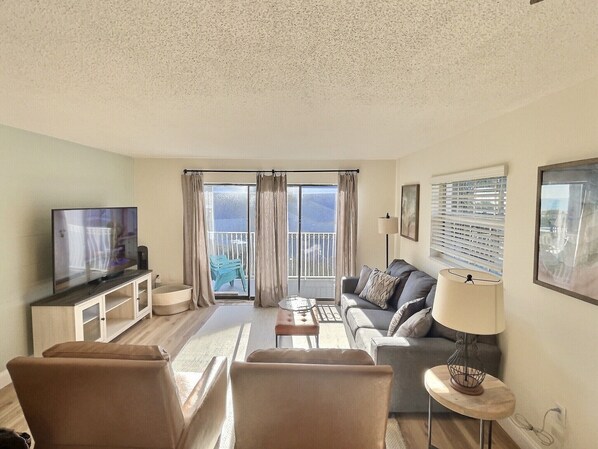 A living room with a television, sofa, and a balcony with curtains. - Ocean View, Family friendly condo at Ola Grande (Cocoa Beach)
