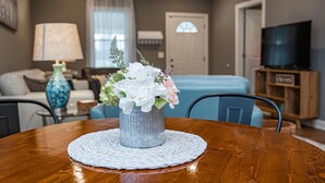 A dining table with a floral centerpiece, a lamp, and a blue vase in a living room with a sofa, TV, and a door in the background. - Cheerful Pet-Friendly Bungalow 1 Block from Downtown Laurens (Laurens)