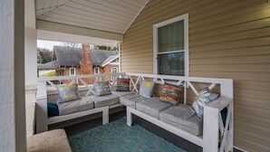 A covered porch with a white bench and cushions, a window, and a view of a brick building. - Cheerful Pet-Friendly Bungalow 1 Block from Downtown Laurens (Laurens)