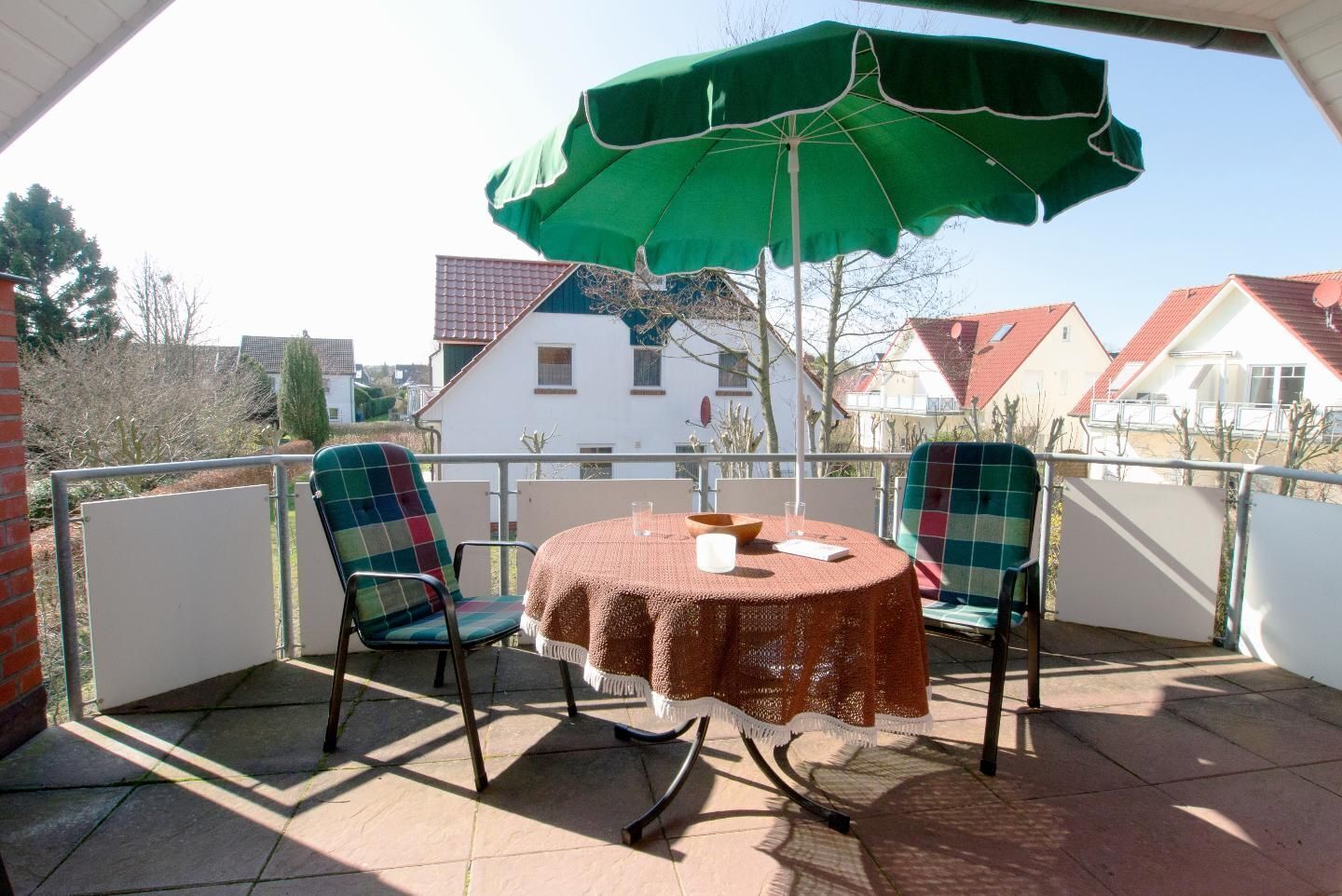 Een balkon met twee stoelen en een tafel, een groene parasol, en een uitzicht op huizen op de achtergrond.