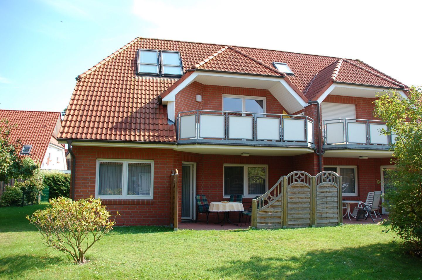Une maison à deux étages, avec un toit recouvert de tuiles rouges, un balcon et un petit jardin.