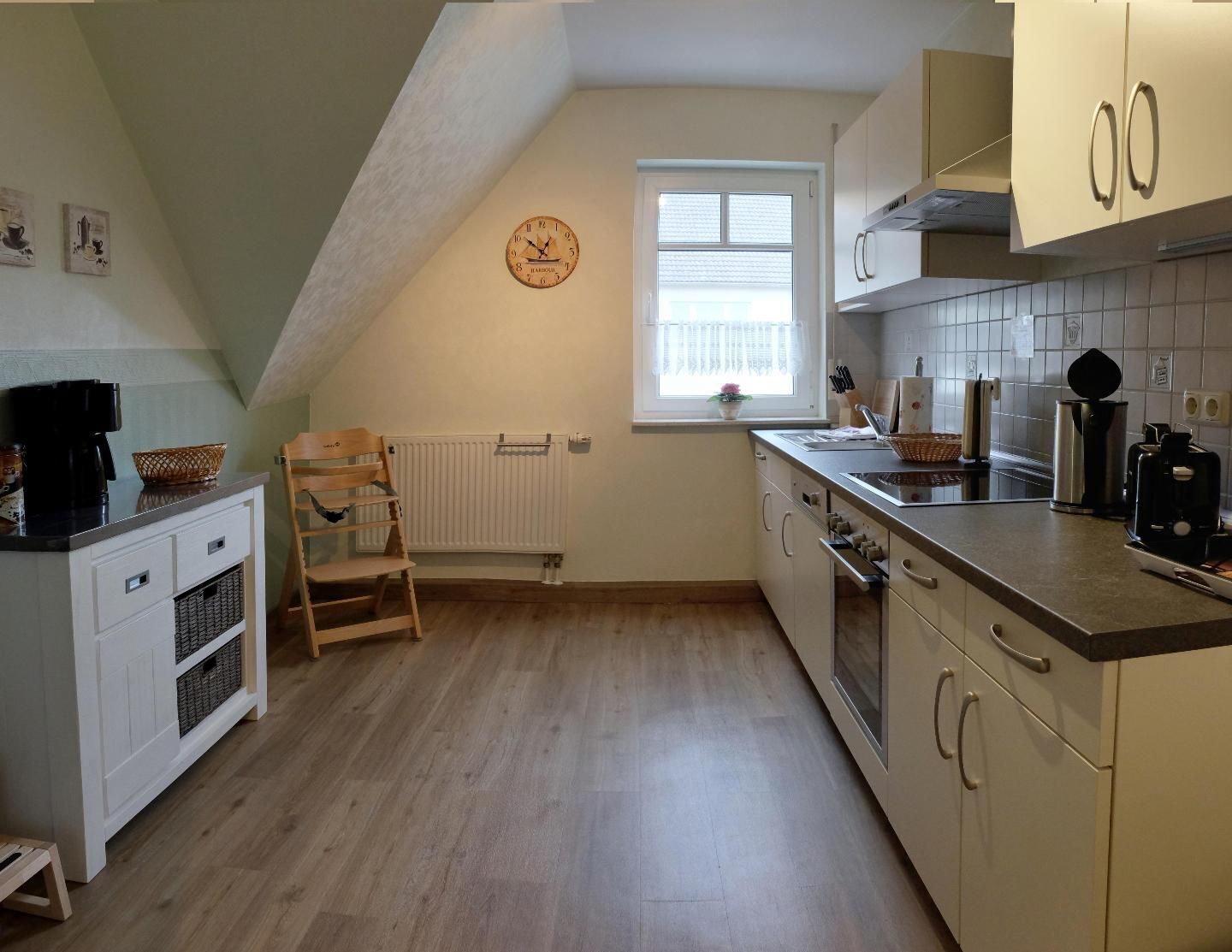 A compact kitchen with a wooden floor, white cabinetry, a clock on the wall, and a window with blinds.