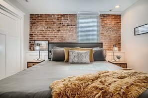A bedroom with a brick wall, a large bed with multiple pillows, a bedside table with a lamp, and a window with blinds.