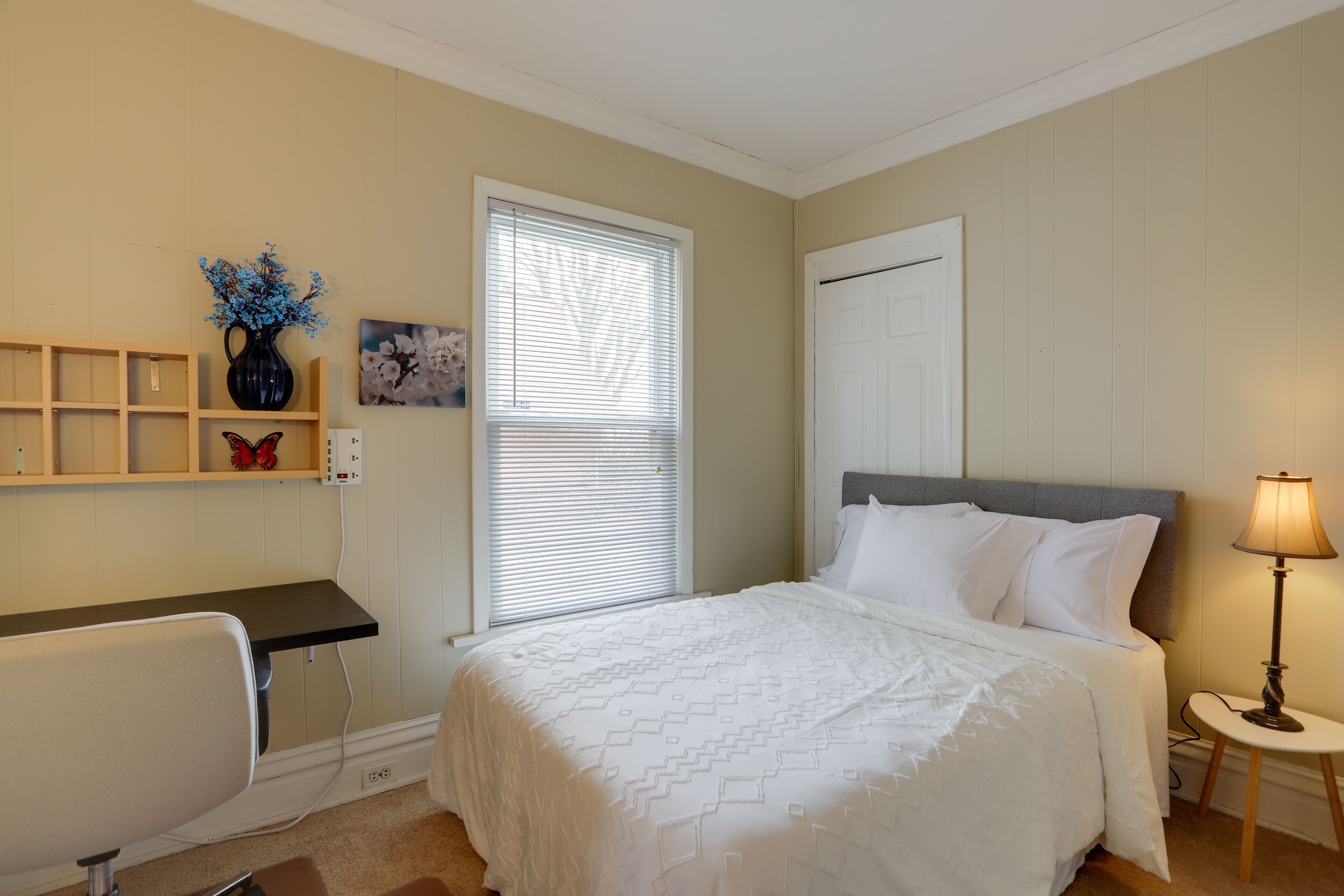 Een slaapkamer met een bed, een bureau, een stoel, een lamp en een raam met jaloezieën.