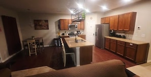 Cocina compacta con armarios de madera, microondas, refrigerador y un pequeño comedor con mesa y sillas.