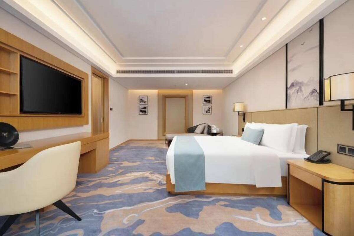 Una habitación de hotel con cama, escritorio, televisor y lámpara de pared.