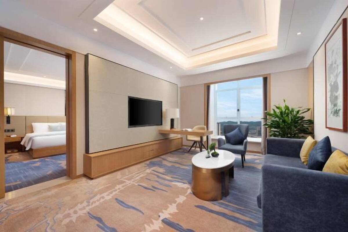 Una habitación de hotel moderna con una cama grande, un televisor de pantalla plana, una zona de estar con sofá y sillas, una mesita pequeña y un ventanal con vistas.