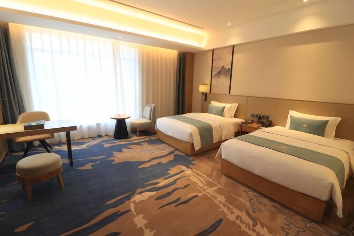 A hotel room with two beds, a desk, a chair, and a large window with curtains.
