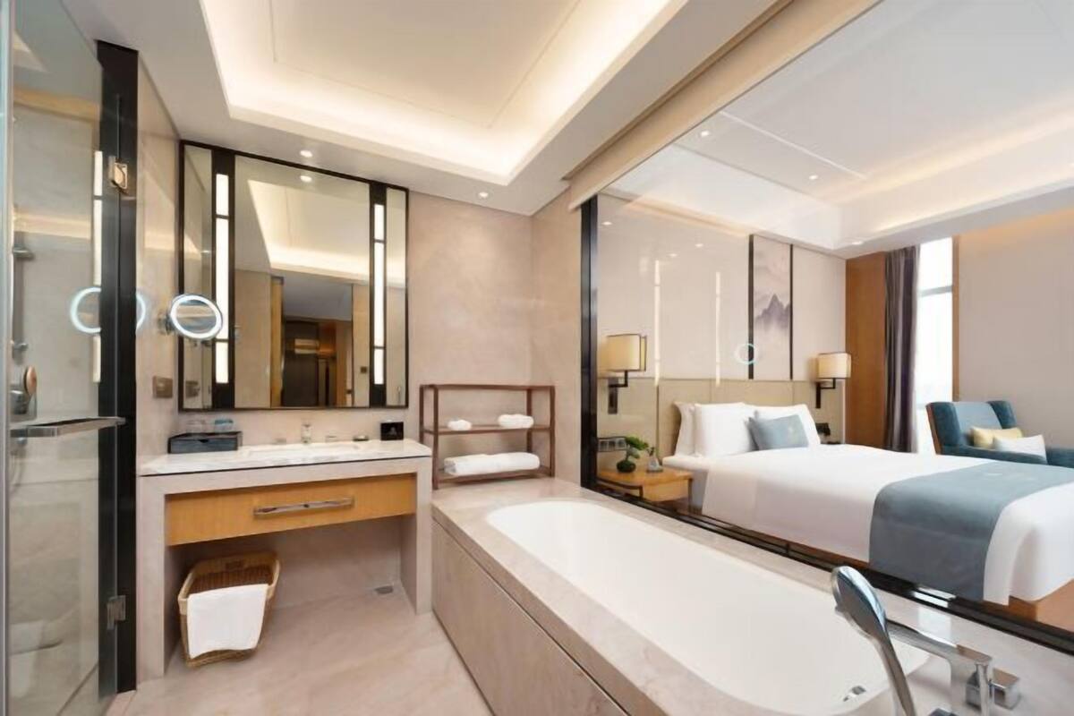 Una habitación de hotel moderna con un espejo grande, una bañera, una cama con una manta azul y una mesita de noche con una lámpara.