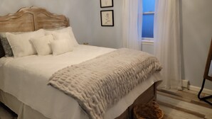 A bedroom with a wooden headboard, a large bed with white bedding, a window with white curtains, and two framed pictures on the wall.