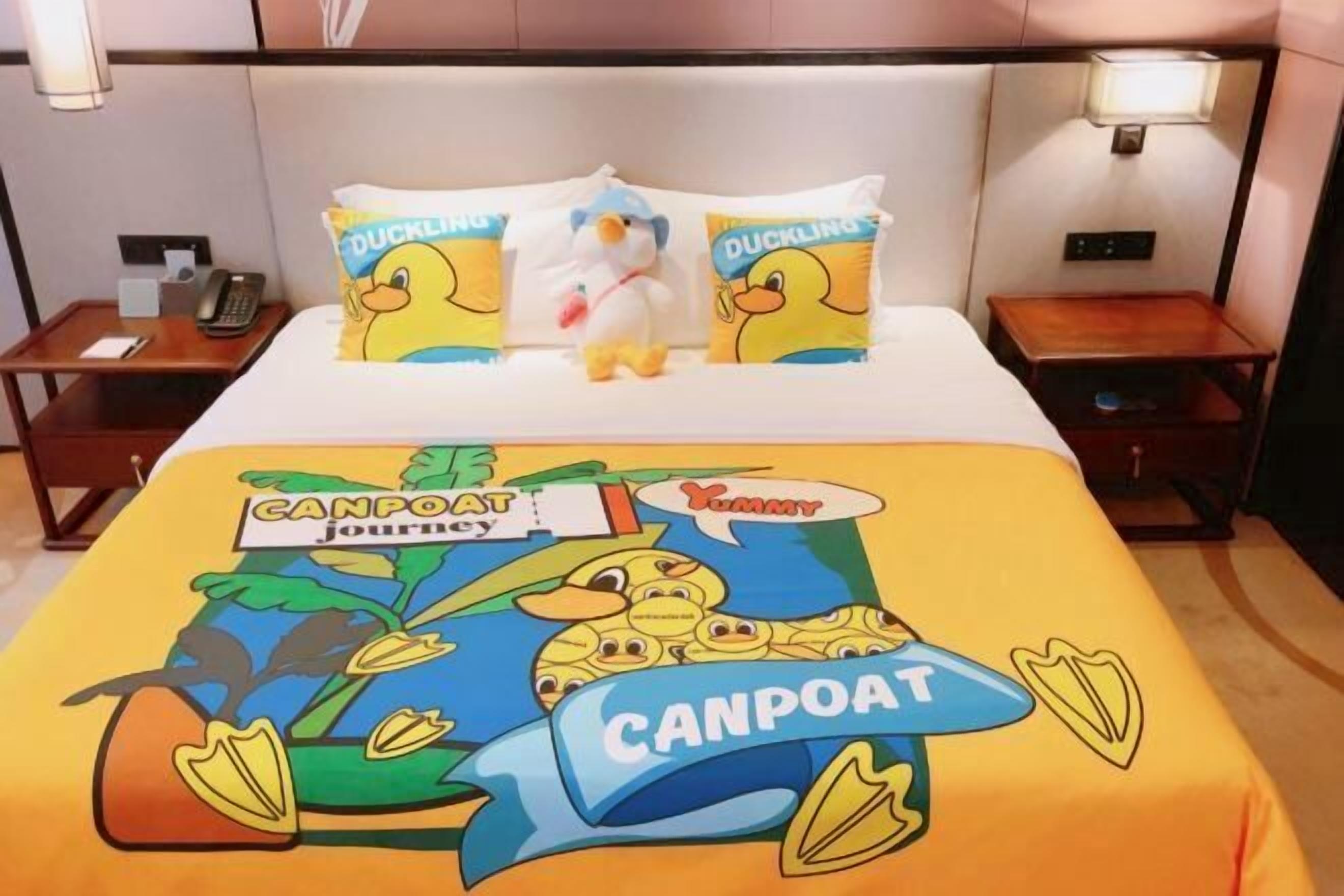 Une chambre d’hôtel avec un lit agrémenté d’une couvre-lit sur le thème de « CANPOAT JOURNEY », deux oreillers décorés de motifs de canetons, une table de chevet avec une lampe et un téléphone sur la table de nuit.