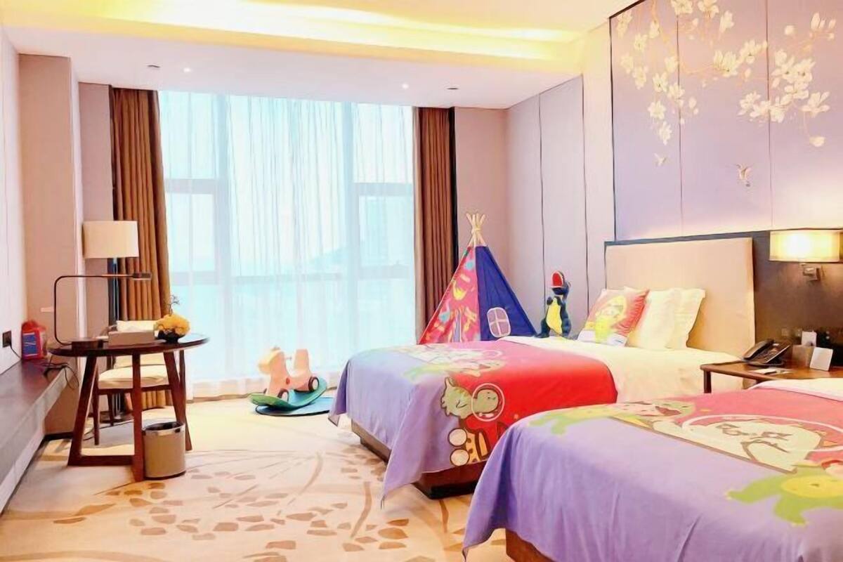Une chambre d’hôtel avec deux lits, un bureau, une chaise, une lampe, une fenêtre avec des rideaux et un mur recouvert d’un papier peint à motifs floraux.