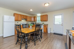 A compact kitchen with wooden cabinets, a white refrigerator, a dining table with chairs, and a window with blinds.