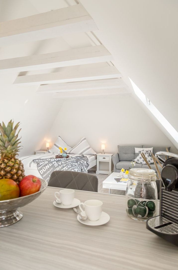 Un dormitorio estilo loft acogedor con techo inclinado, una cama con ropa de cama estampada, un sofá gris y una mesa de comedor con tazas y un frutero.