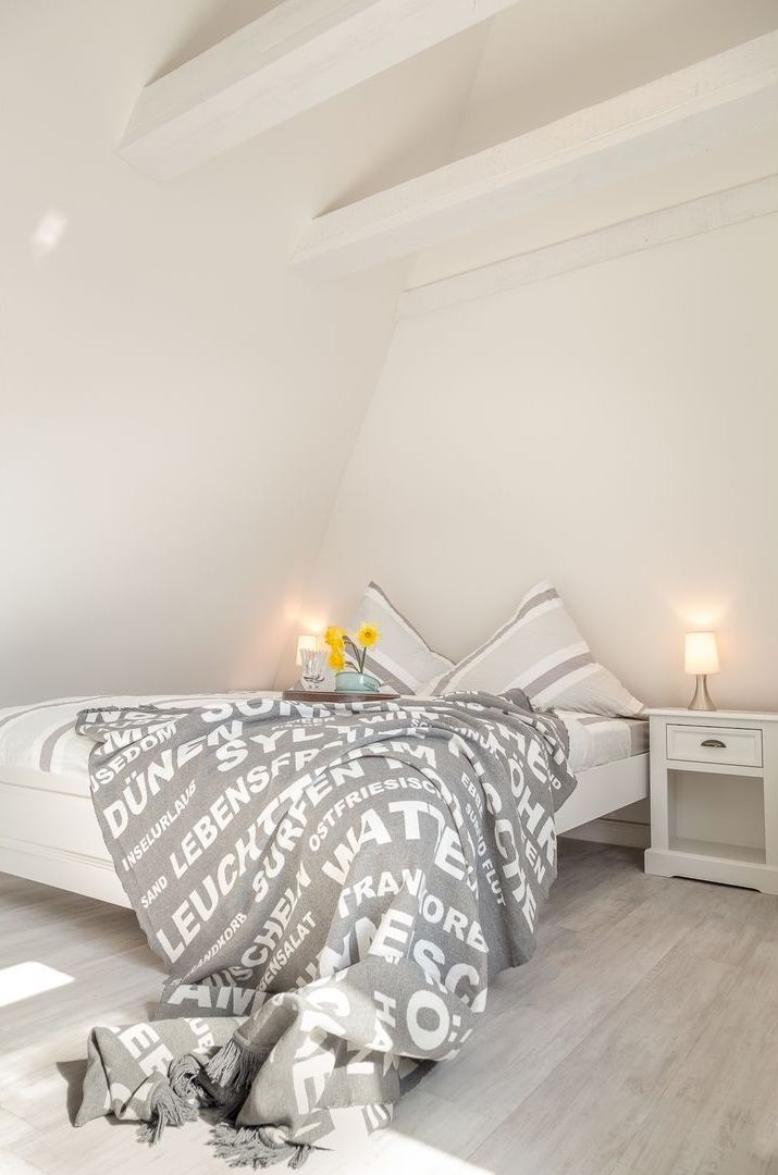 Un dormitorio con una cama cubierta con una manta que tiene texto, una mesita de noche con una lámpara y un pequeño jarrón con flores.