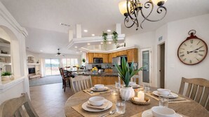 A spacious dining area with a round table set for four, surrounded by wooden chairs. A kitchen with wooden cabinets and a central island is visible in the background.