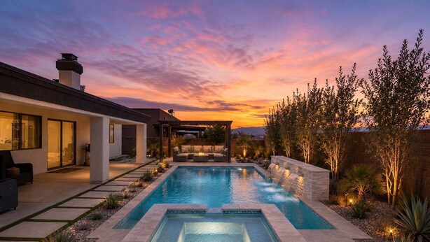 A modern outdoor pool with a waterfall feature, surrounded by a landscaped garden and a house with a covered patio.