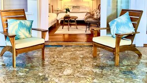 A room with a bed, two wooden chairs with cushions, a small table, and a floor with a stone pattern. - Beautiful Beachside Bungalow (Kailua)