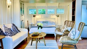 A spacious living room with a white sofa, wicker chairs, a round coffee table, and a large bed with a view of greenery outside. - Beautiful Beachside Bungalow (Kailua)