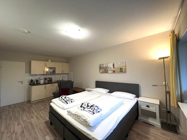 Habitación de hotel con cama, mesitas de noche, escritorio, silla, kitchenette y un cuadro en la pared.