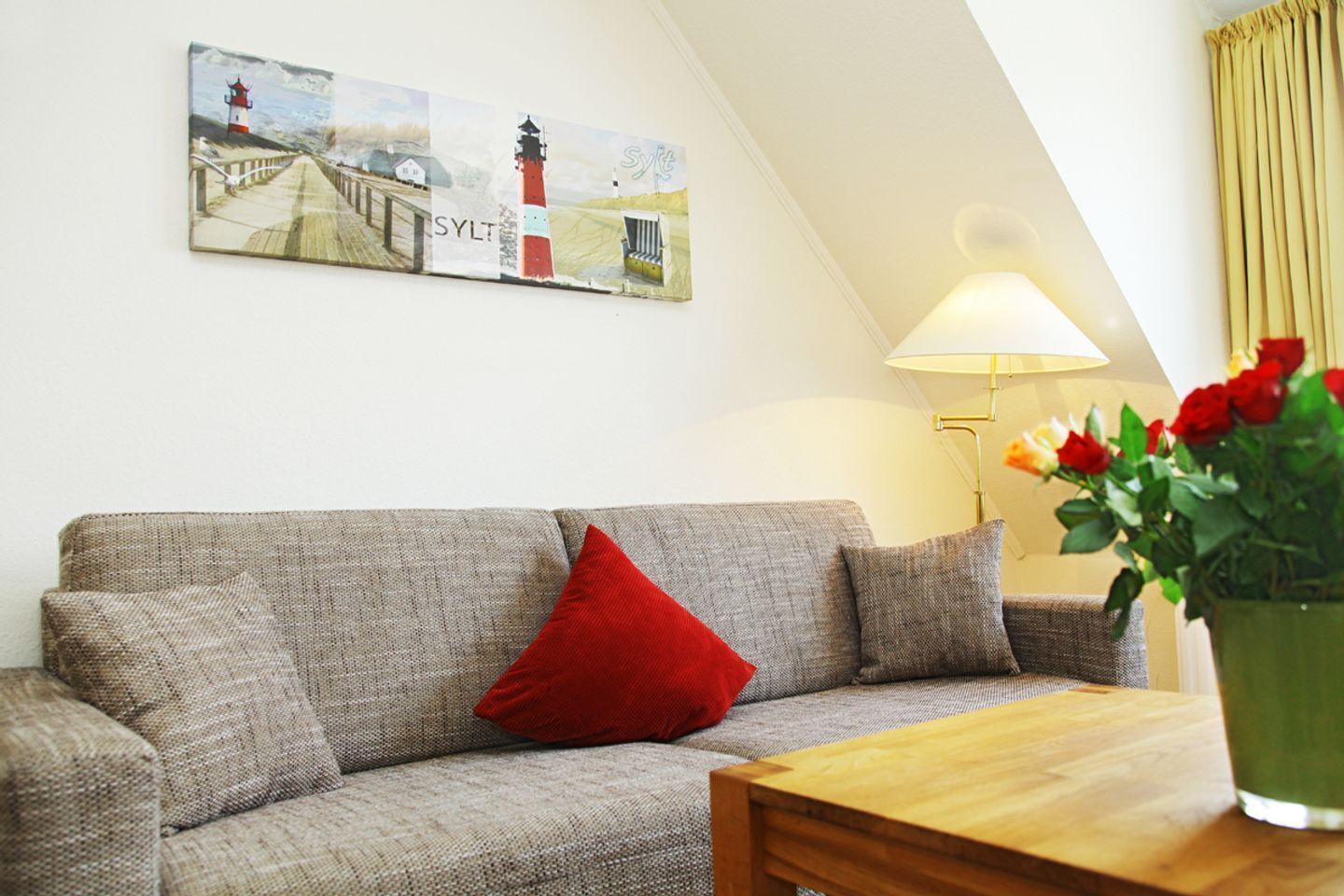 Ein gemütliches Wohnzimmer mit einem grauen Sofa, einem roten Kissen, einem Holztisch und einem Gemälde eines Leuchtturms.