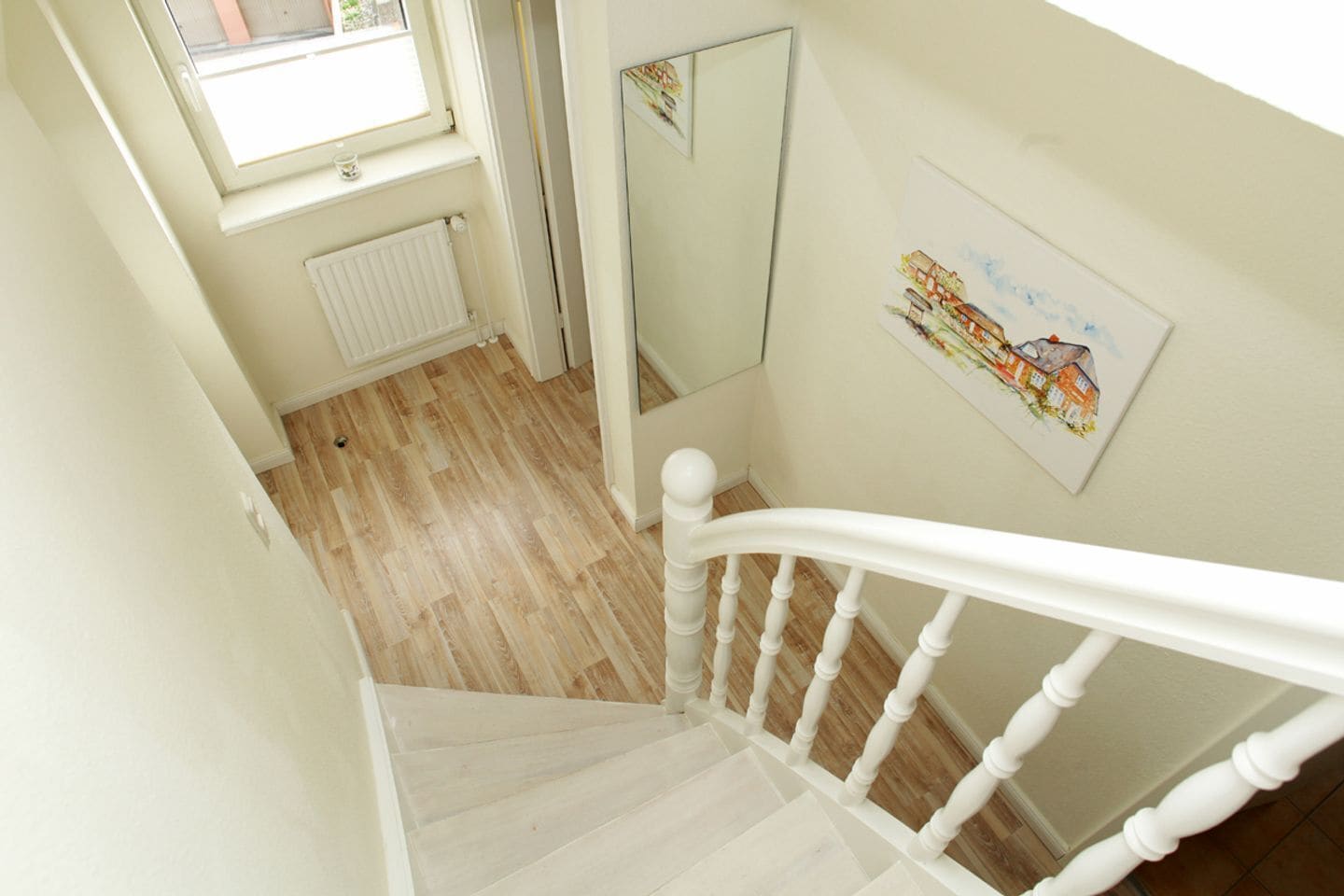 A staircase with white balusters leading to a landing. A radiator is visible on the wall below the window, and a framed picture hangs on the wall.