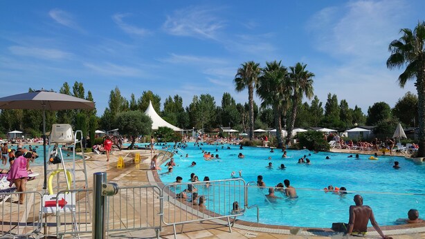 A large outdoor swimming pool surrounded by palm trees and umbrellas, with people swimming and sunbathing. - Mobile Home (Lattes)