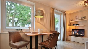 Una sala de estar moderna con una mesa de comedor de madera, dos sillas marrones, un televisor y un ventanal con vistas a árboles.