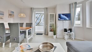 A modern living room with a dining area, a TV mounted on the wall, and a view of the outdoors through large windows.