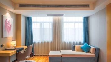 A modern hotel room with a desk, chair, sofa, and large windows with curtains.