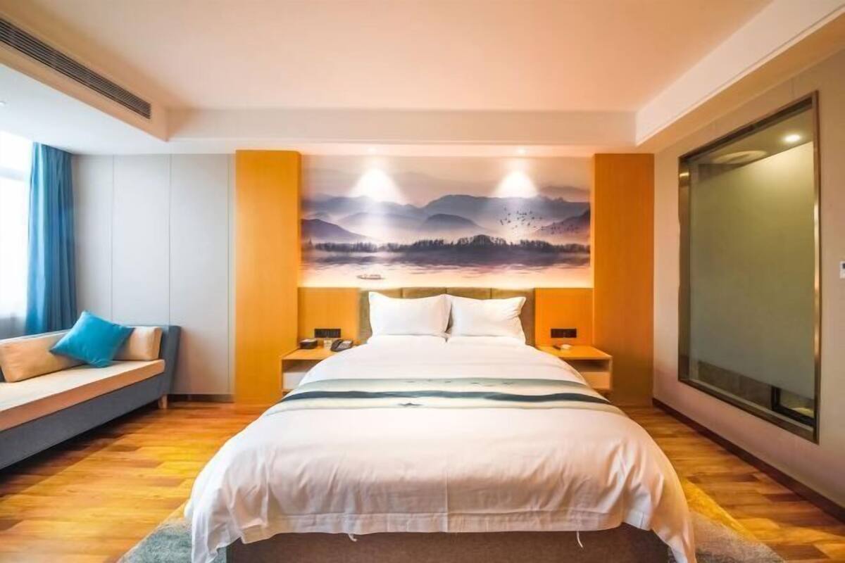 A hotel room with a bed, bedside tables, a sofa, a large window, and a wall-mounted artwork.