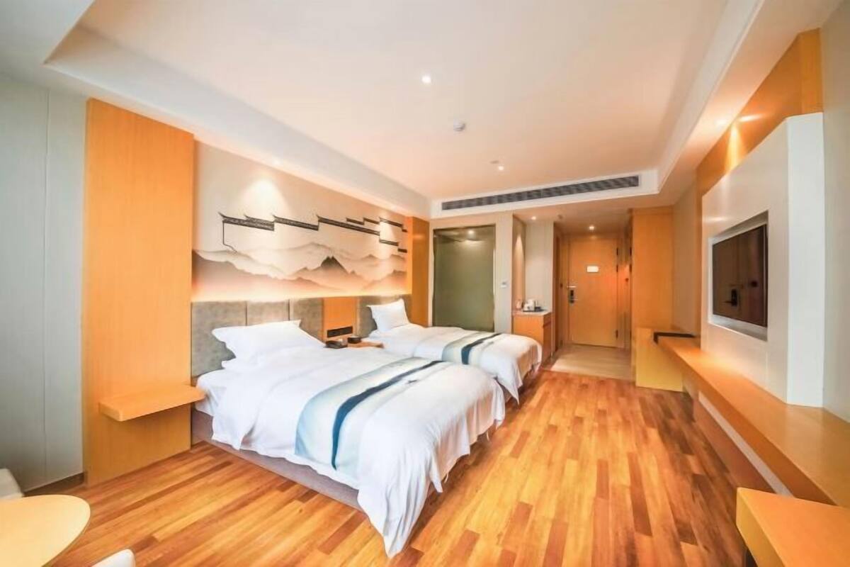 A hotel room with a wooden floor, two beds with white bedding, a wall-mounted TV, and a modern light fixture.