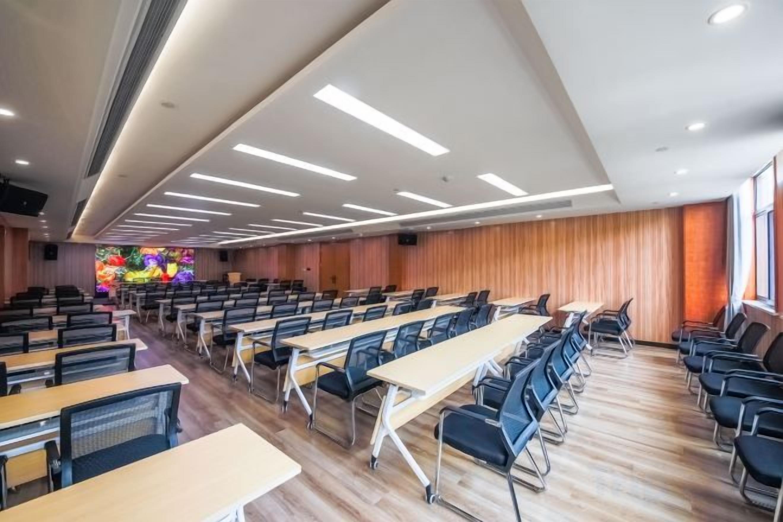 A conference room with rows of tables and chairs, a large screen, and a wooden panel wall.