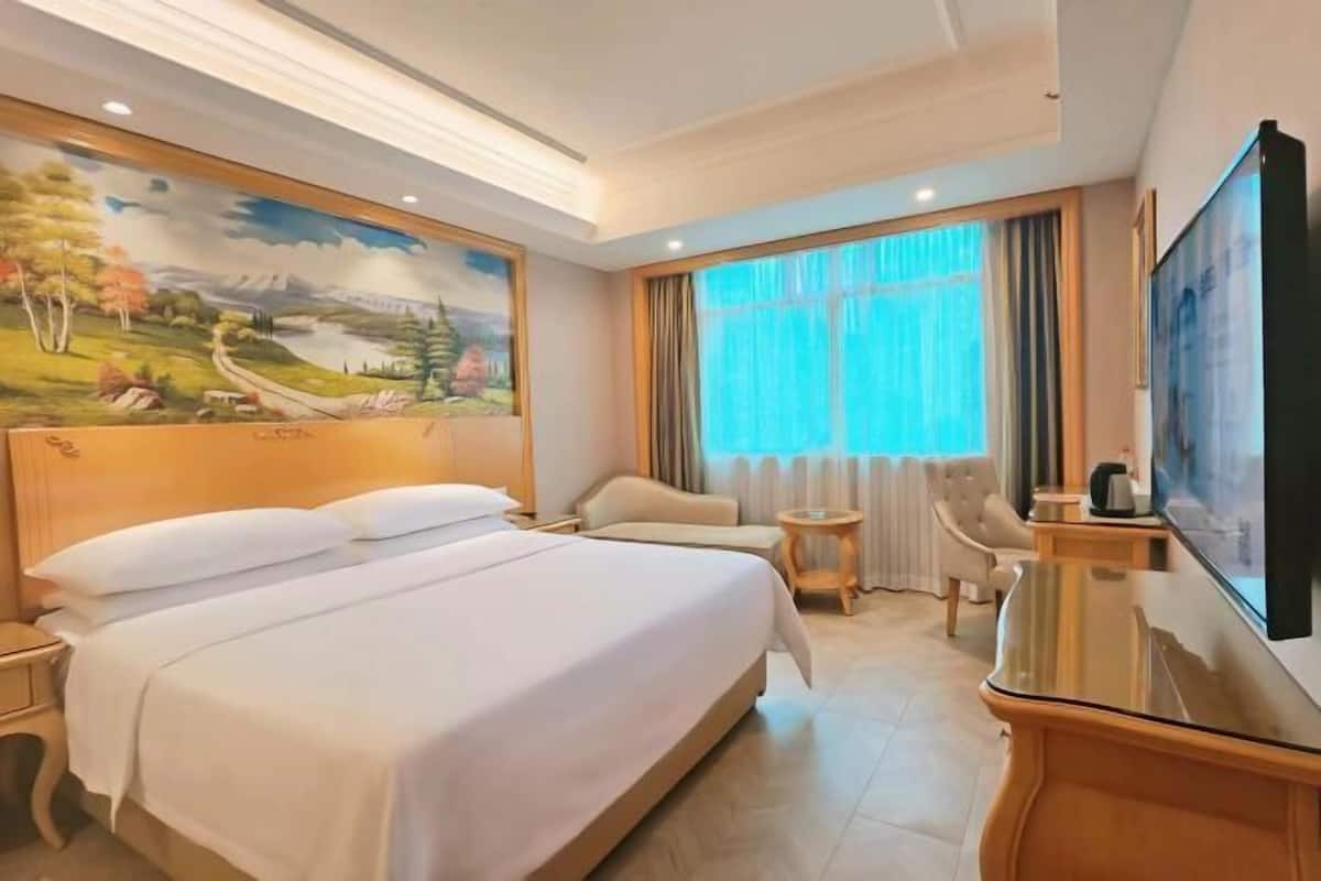 A hotel room with a large bed, a TV, a desk, a chair, and a painting on the wall.