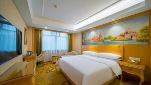 A hotel room with a large bed, a flat-screen TV, a desk with a chair, a small round table, and a painting of a historical building on the wall. - Vienna Hotel (Jishou Qianzhou Ancient City Branch) (Jishou)