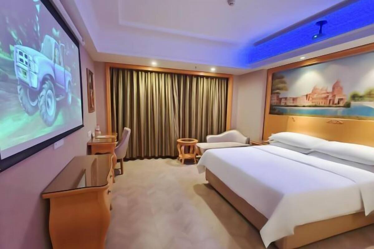 A hotel room with a bed, a desk, a chair, a small table, a projector screen, and a curtain.