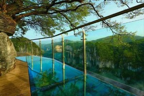Un pasillo con fondo de cristal que lleva a una piscina con vistas a una cadena montañosa cubierta de bosque.