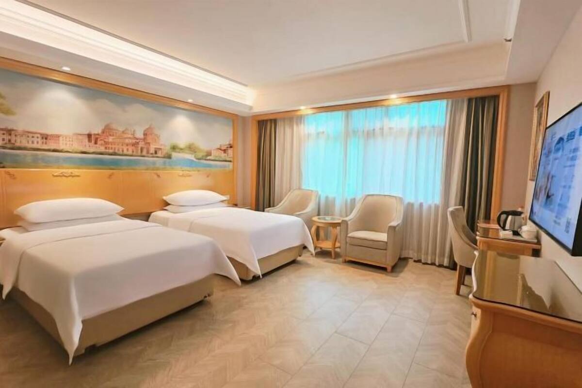 A hotel room with two beds, a desk, a chair, a television, and a painting on the wall.